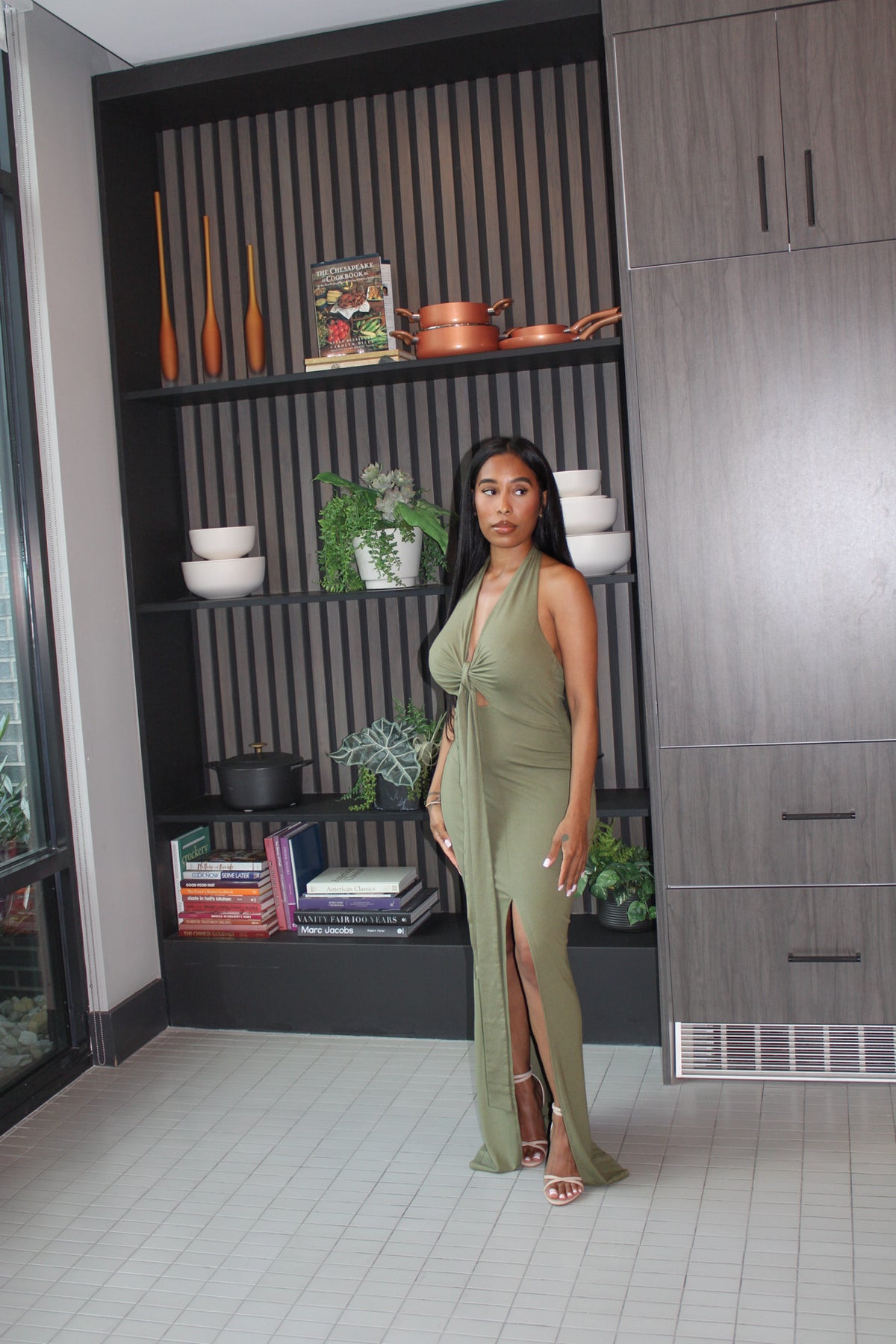 A woman wearing a sage green halter-neck maxi dress with a thigh-high slit, standing in a modern interior room with a slatted wood display wall and cabinets behind her.